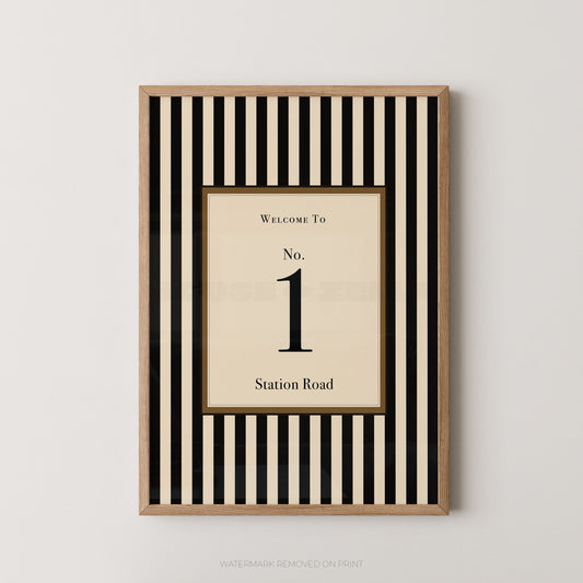 A framed vertical wall art print with alternating black and cream stripes, featuring a central cream panel that reads 'WELCOME TO No. 1 Station Road' in black serif text, shown against a light gray wall.