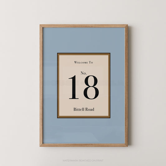 Framed wall art print with a light blue background and a central cream panel reading 'Welcome To No. 18 Bittell Road' in black text, shown in a light wood frame against a white wall.