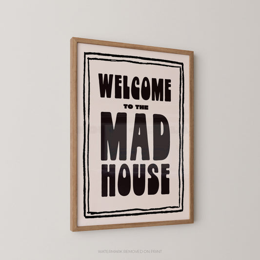 Framed wall art print with the text 'WELCOME TO THE MAD HOUSE' in bold black lettering on a cream background, enclosed in a light wood frame and shown hanging on a light gray wall.