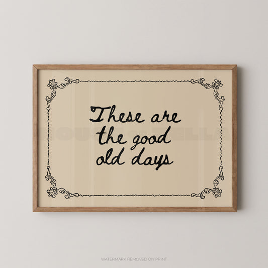 Framed wall art print with the phrase “These are the good old days” in black script on a warm cream background, bordered by a light wood frame and small decorative corner flourishes.