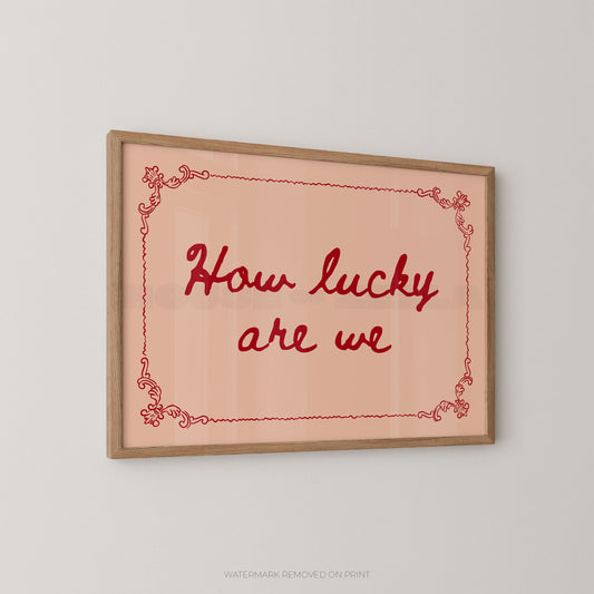 Framed wall art print with the phrase “How lucky are we” in red script on a pale peach background, bordered by a thin red decorative line and a light wood frame, shown against a light gray wall.