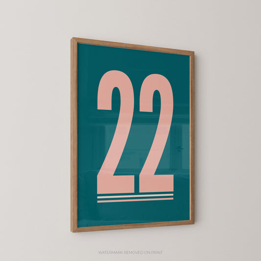 Framed wall art print featuring the number 22 in pale pink on a teal background, shown hanging on a light gray wall.