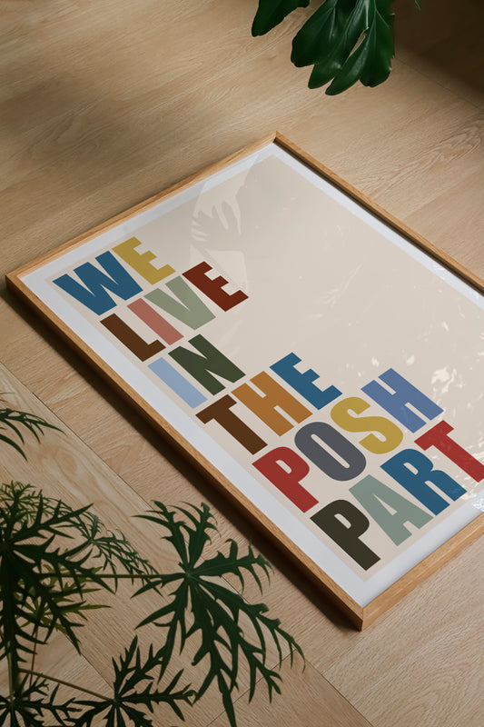 A framed typographic wall art print reading “WE LIVE IN THE POSH PART” in multicolored letters on a cream background, shown at an angle on a light wood floor with green plants around it.