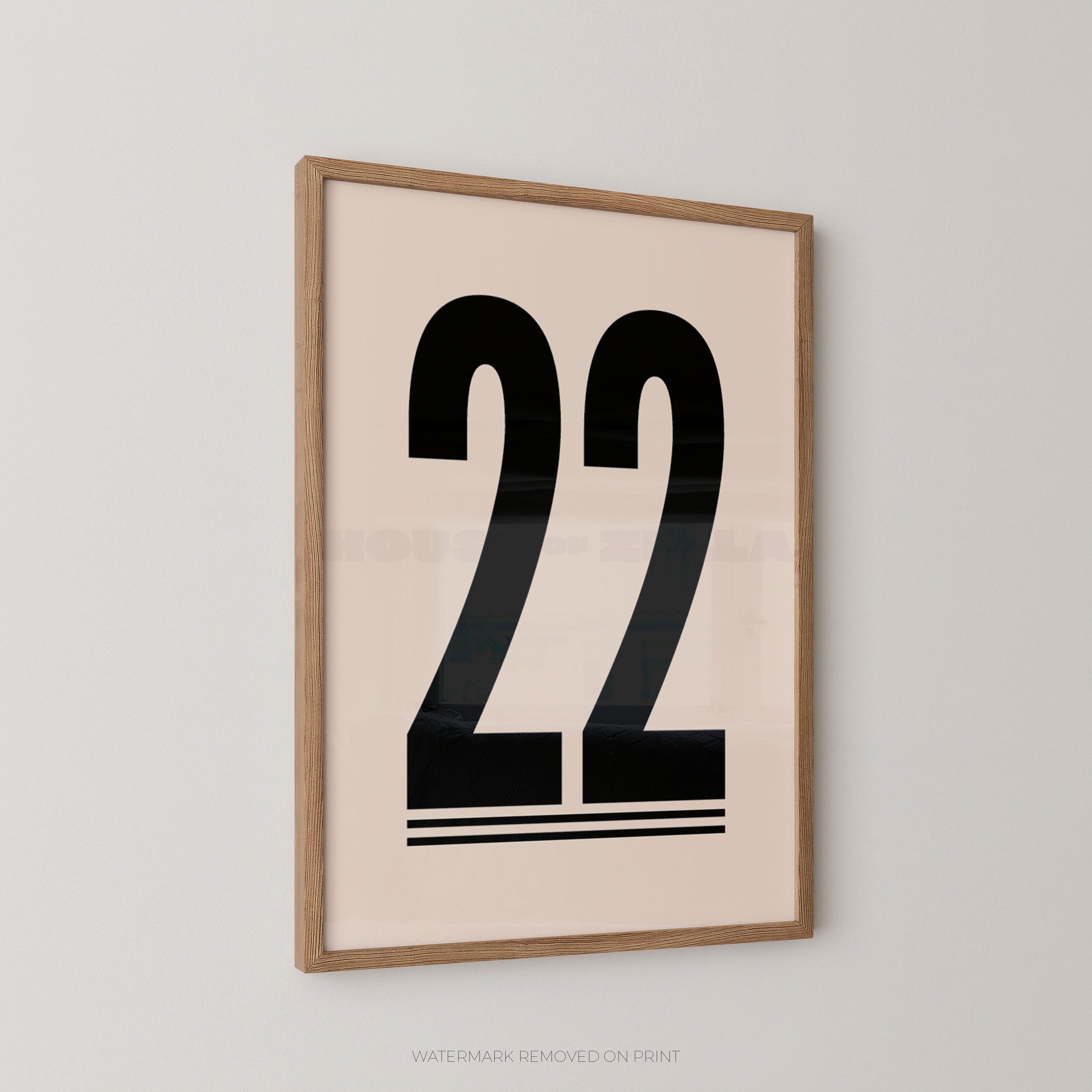 A framed minimalist art print featuring the black number 22 on a warm cream background, enclosed in a light natural wood frame and mounted on a neutral light gray wall.