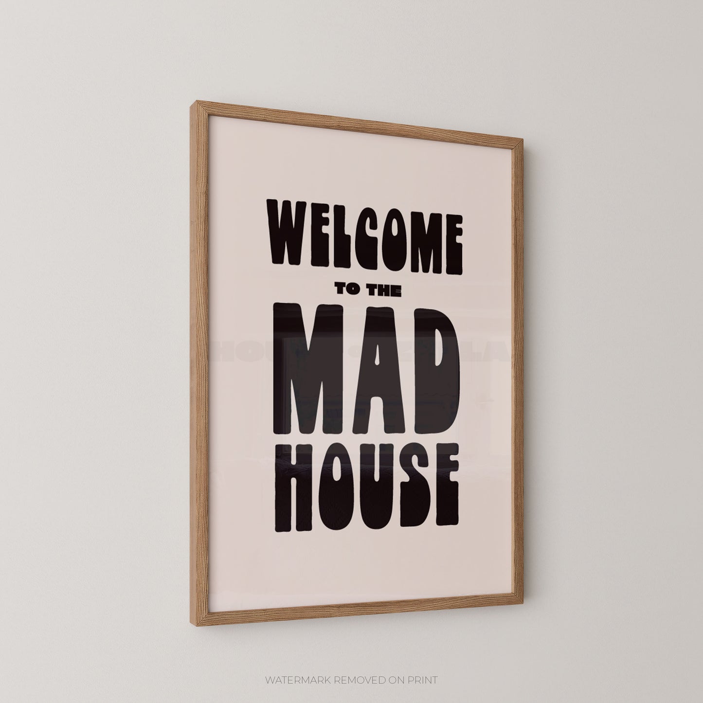 A framed typographic wall art print reading “WELCOME TO THE MAD HOUSE” in bold black lettering on a pale blush background, shown hanging on a light gray wall.