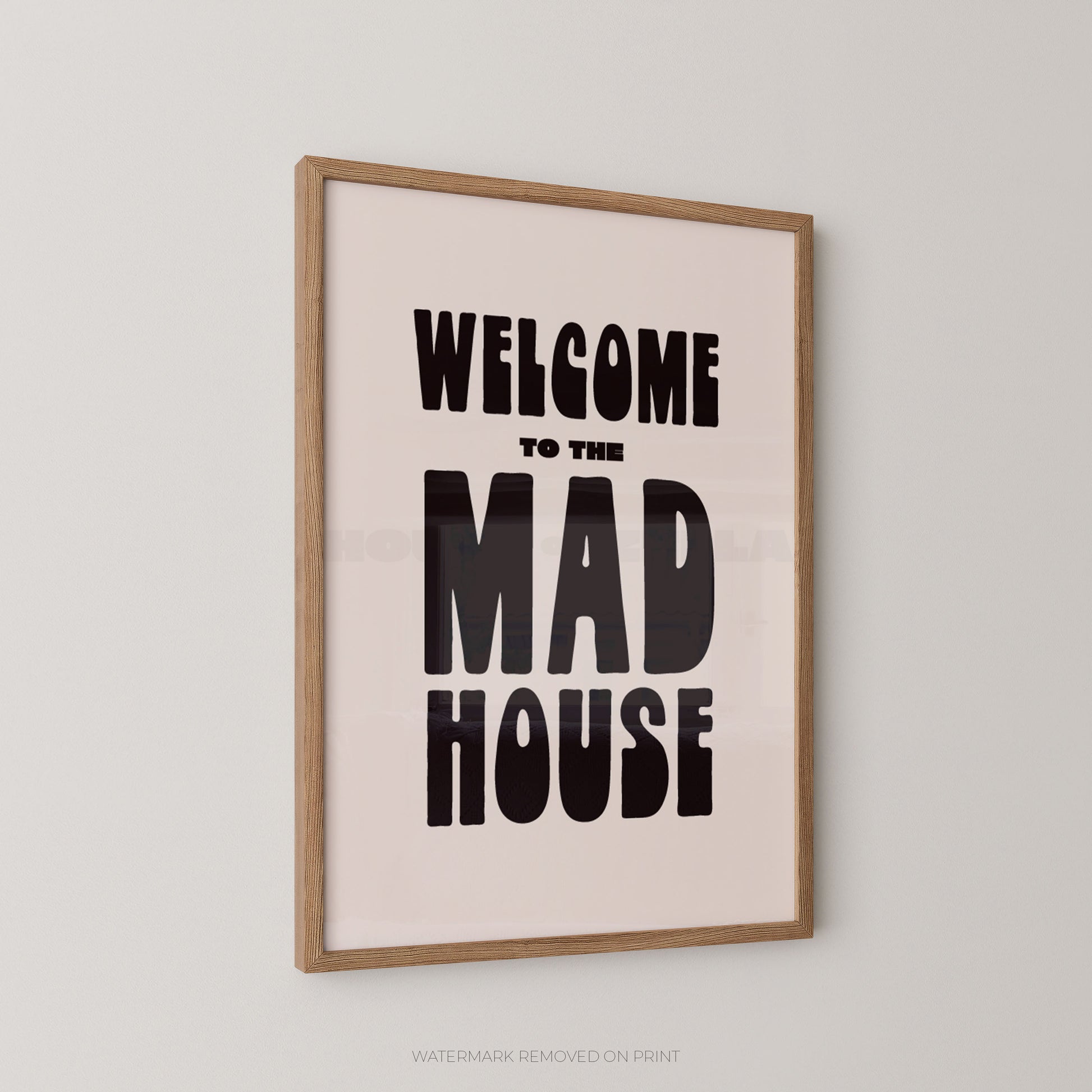 A framed typographic wall art print reading “WELCOME TO THE MAD HOUSE” in bold black lettering on a pale blush background, shown hanging on a light gray wall.