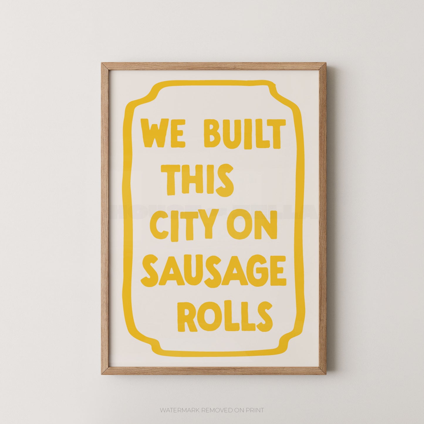A framed typographic wall art print with the phrase “WE BUILT THIS CITY ON SAUSAGE ROLLS” in yellow text inside a yellow outlined badge shape, shown against a light neutral wall.