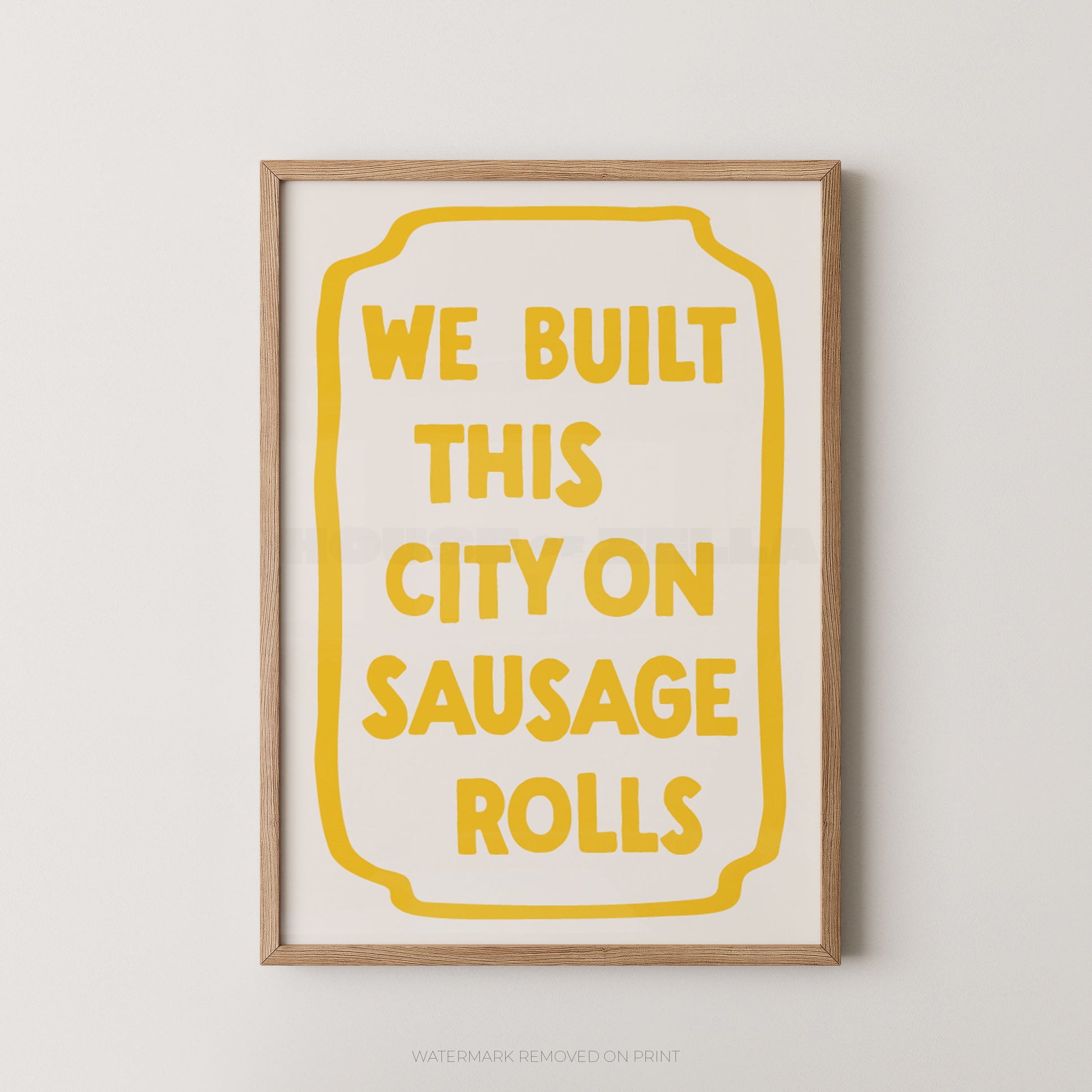 A framed typographic wall art print with the phrase “WE BUILT THIS CITY ON SAUSAGE ROLLS” in yellow text inside a yellow outlined badge shape, shown against a light neutral wall.