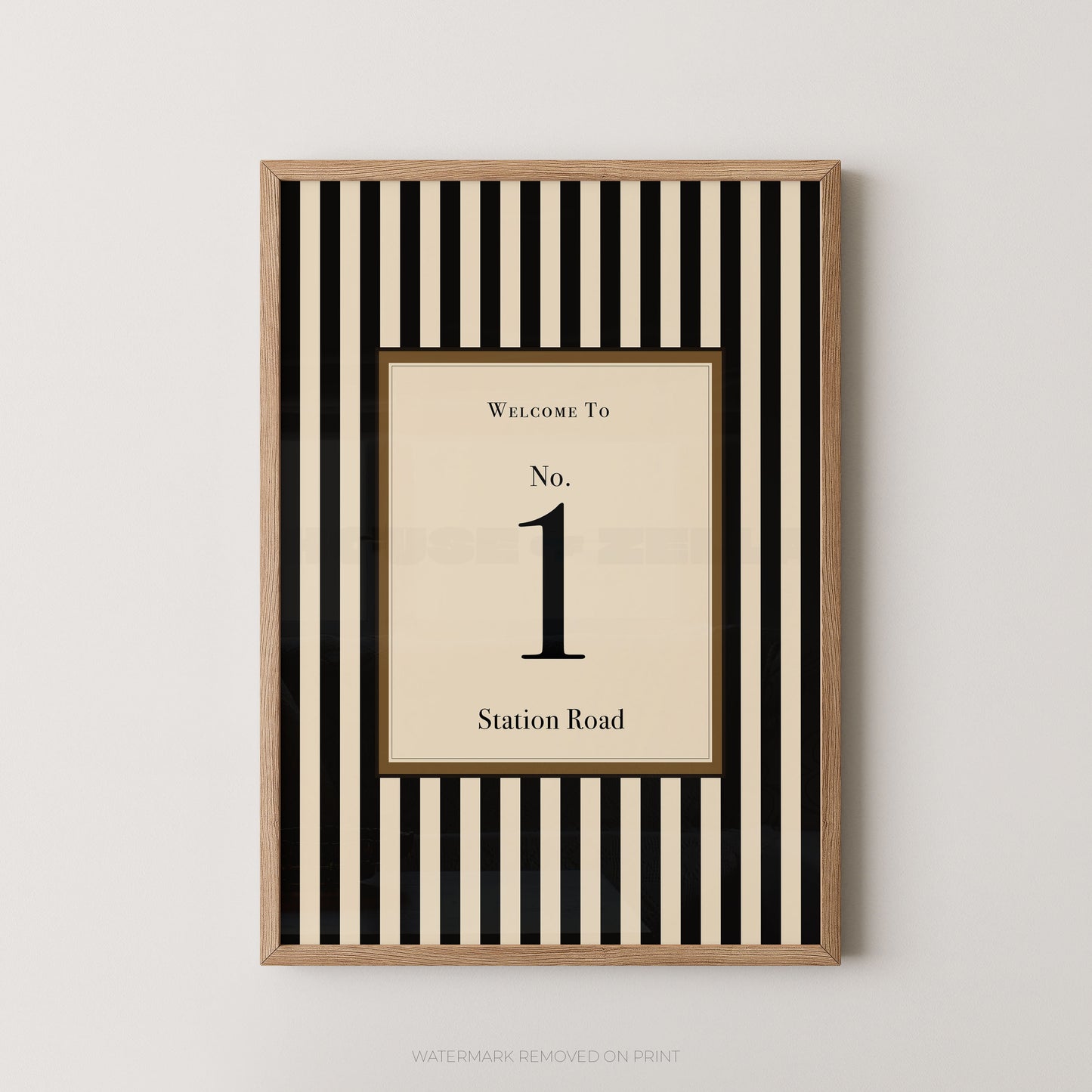 A framed vertical wall art print with alternating black and cream stripes, featuring a central cream panel that reads 'WELCOME TO No. 1 Station Road' in black serif text, shown against a light gray wall.