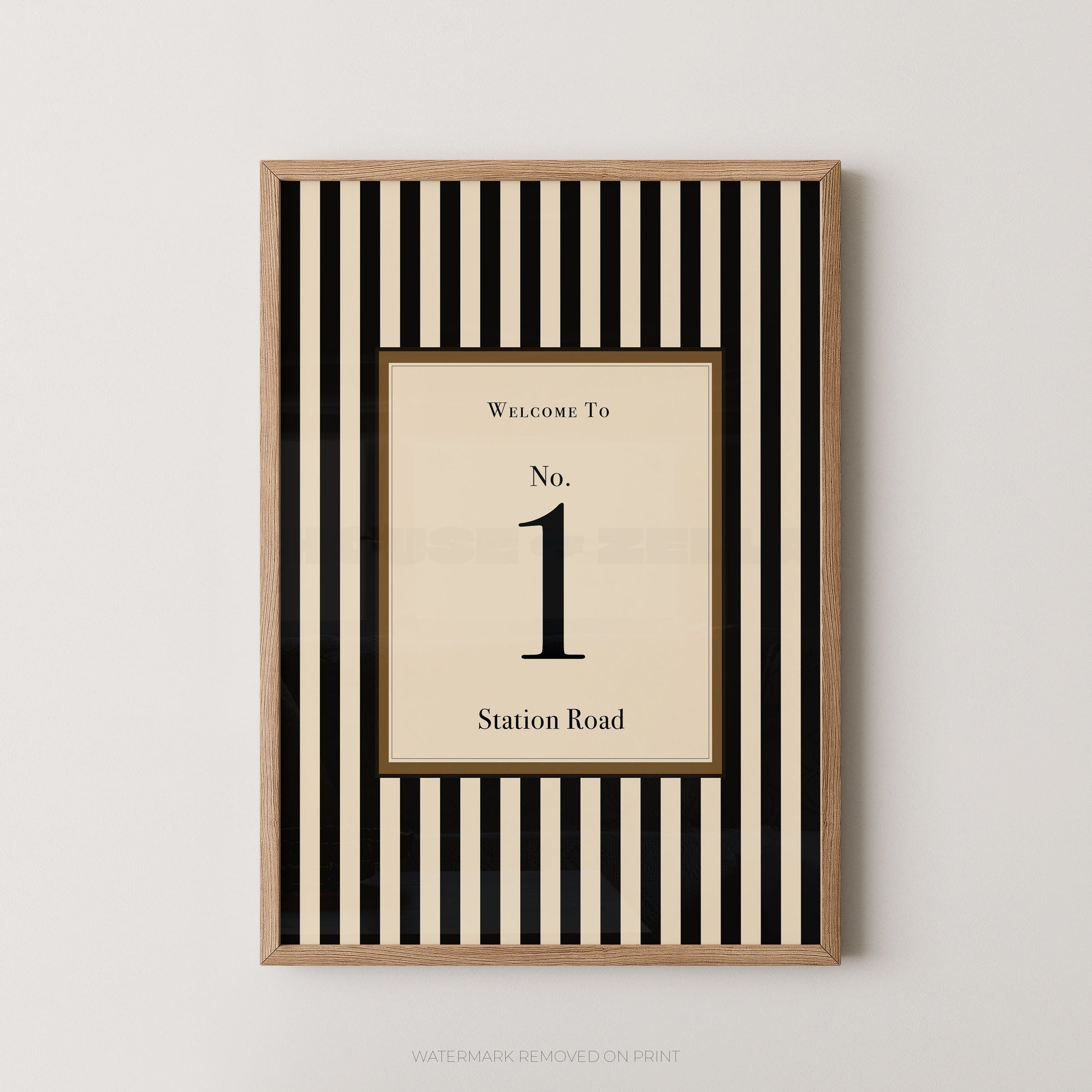 A framed vertical wall art print with alternating black and cream stripes, featuring a central cream panel that reads 'WELCOME TO No. 1 Station Road' in black serif text, shown against a light gray wall.