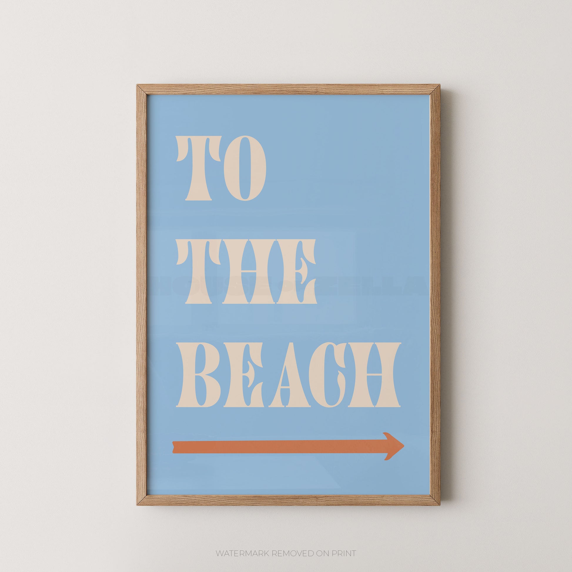 A framed typographic wall art print with a light blue background, cream text reading 'TO THE BEACH,' and a coral-orange arrow pointing right, shown against a light neutral wall.