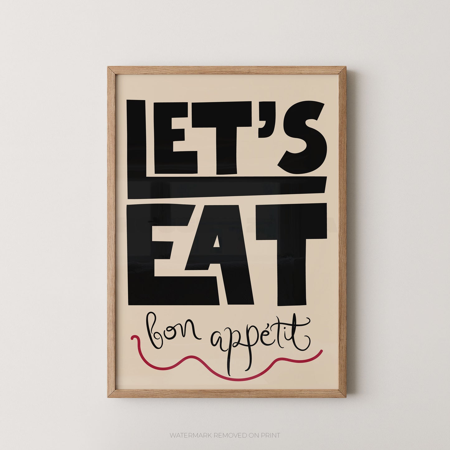 Framed typographic wall art with the words “LET’S EAT” in large black block letters on a warm cream background, with “bon appetit” in black script and a red wavy underline, shown in a light wood frame against a light gray wall.