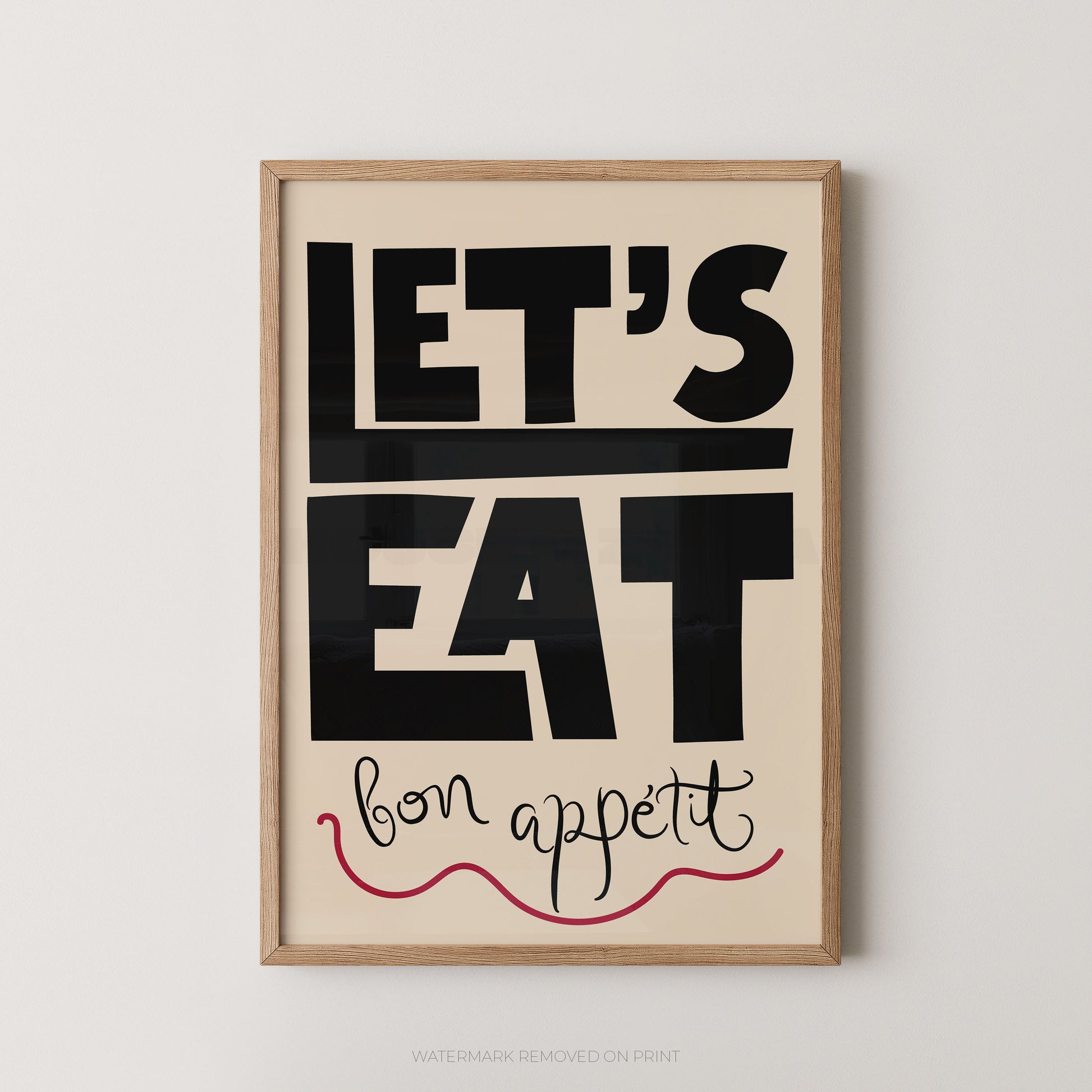 Framed typographic wall art with the words “LET’S EAT” in large black block letters on a warm cream background, with “bon appetit” in black script and a red wavy underline, shown in a light wood frame against a light gray wall.