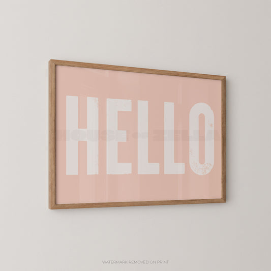Framed wall art print with the word "HELLO" in large white letters on a soft pink background, shown hanging on a light neutral wall.