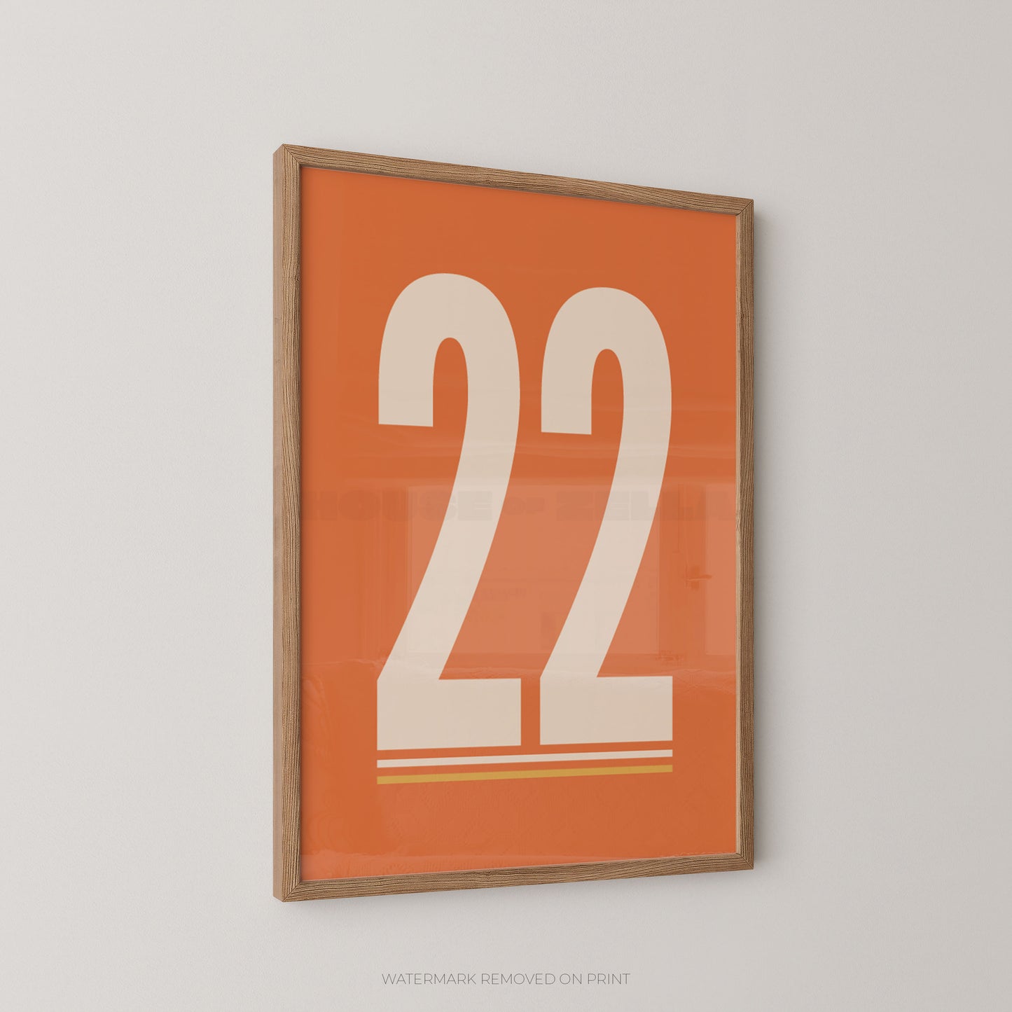 A framed art print featuring the number 22 in bold white typography on a vibrant orange background, enclosed in a light brown wood frame and displayed on a neutral light gray wall.