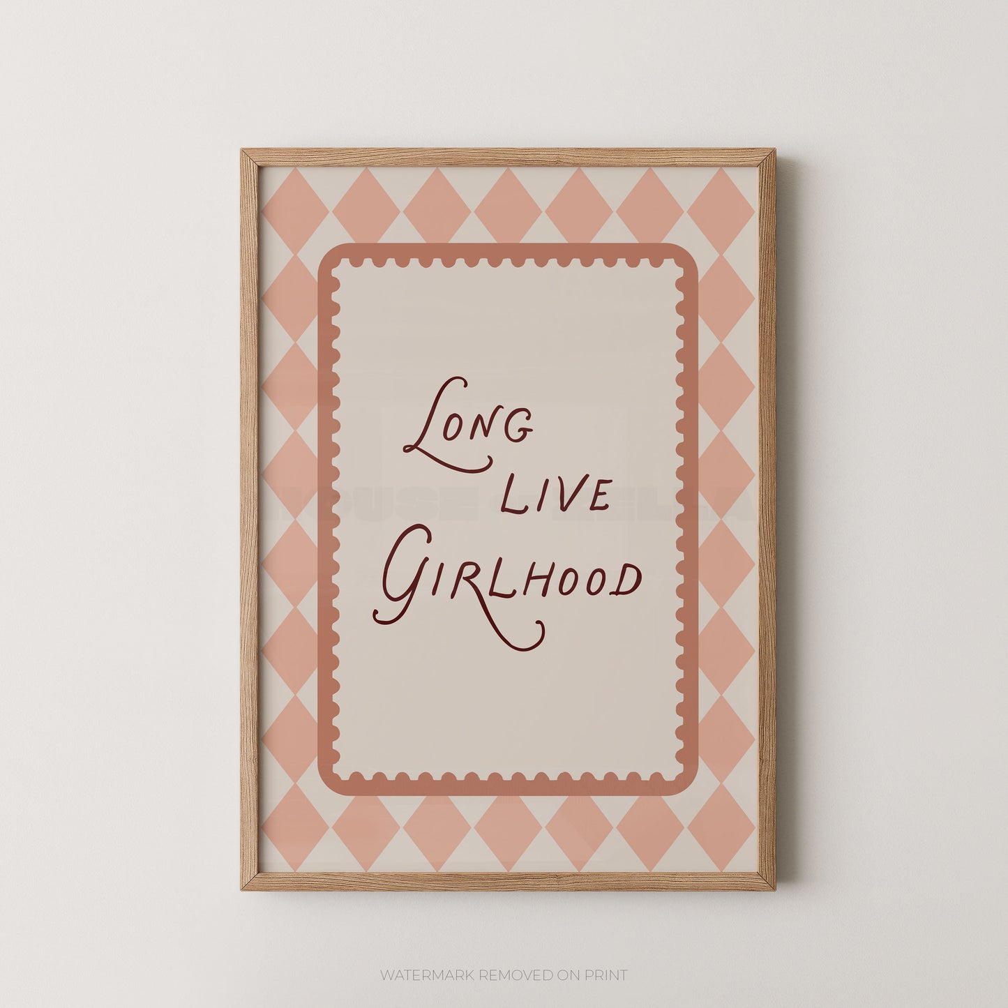 Framed wall art print with the phrase “Long Live Girlhood” in dark script inside a scalloped border, set against a pink and cream diamond pattern. The print is housed in a light natural wood frame and is shown hanging on a neutral light gray wall.