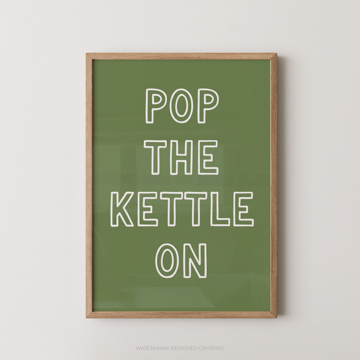 A framed typographic wall art print with the phrase “POP THE KETTLE ON” in white outlined letters on an olive green background, shown hanging against a light neutral wall.