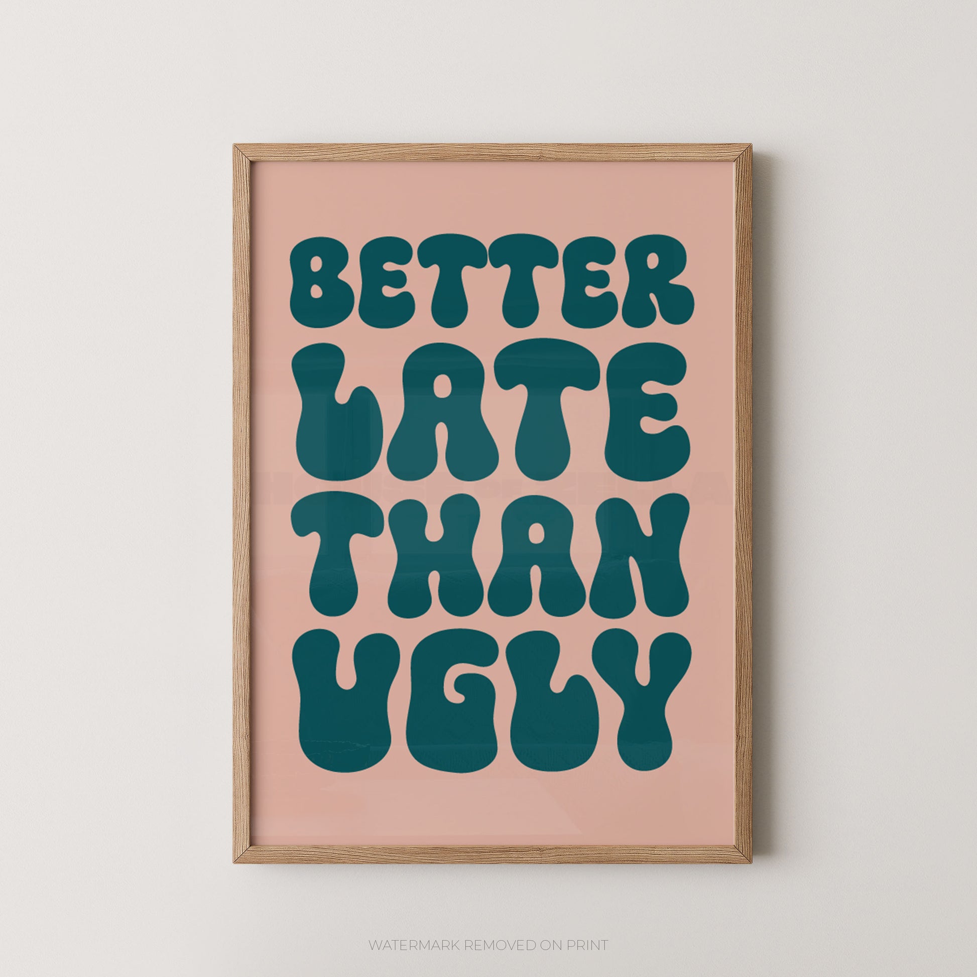 Framed typographic wall art with the phrase “BETTER LATE THAN UGLY” in large teal retro letters on a blush pink background, shown in a light wood frame against a light neutral wall.