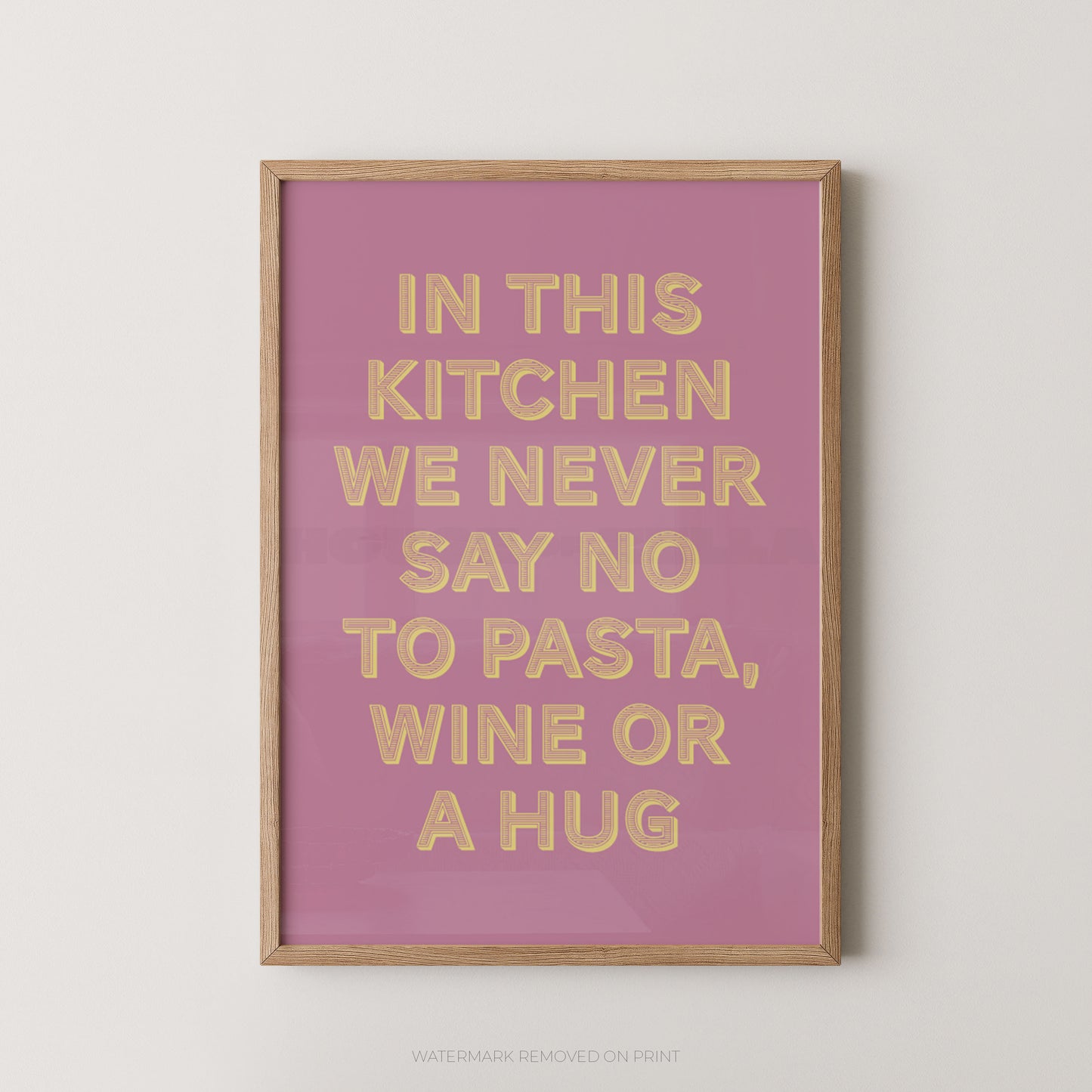 A framed typographic wall art print with a pink background and gold text reading, 'IN THIS KITCHEN WE NEVER SAY NO TO PASTA, WINE OR A HUG,' shown against a light neutral wall.
