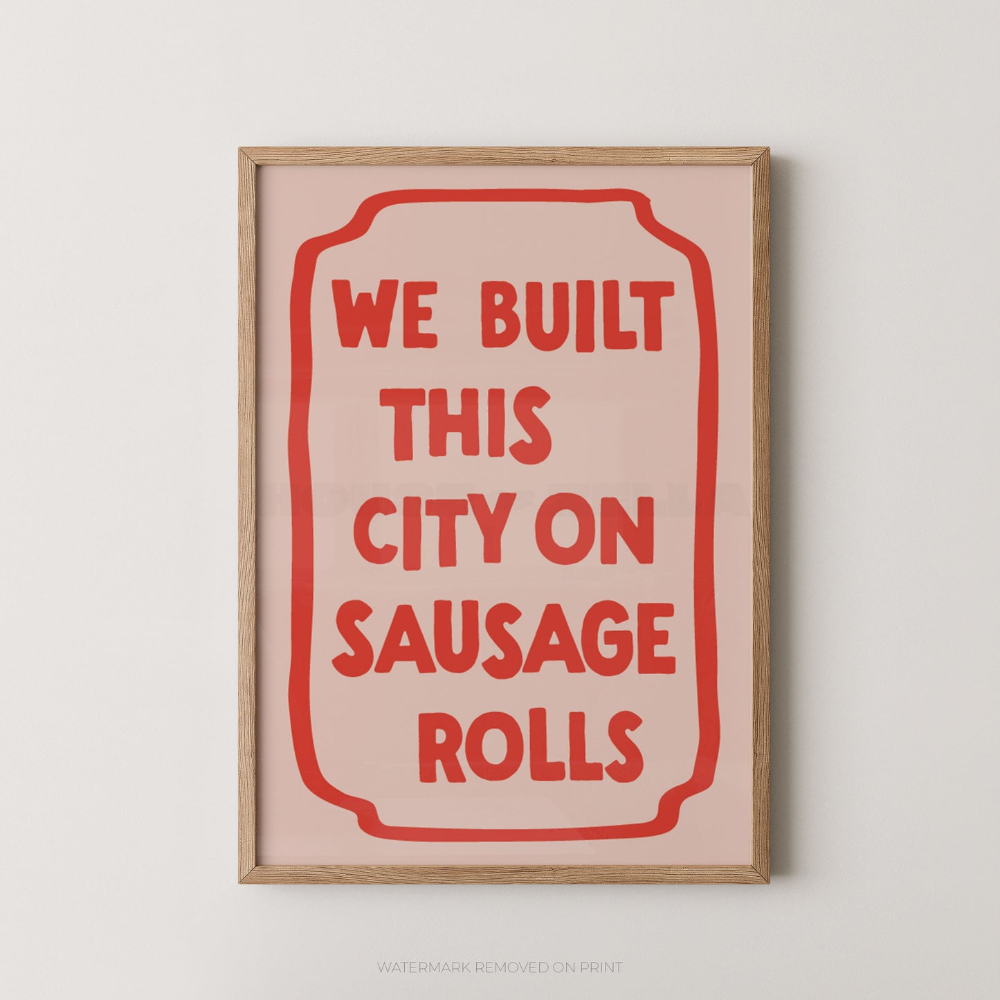 A framed typographic wall art print with a light wood frame, pale pink background, and bold red text reading “WE BUILT THIS CITY ON SAUSAGE ROLLS,” shown hanging against a light neutral wall.