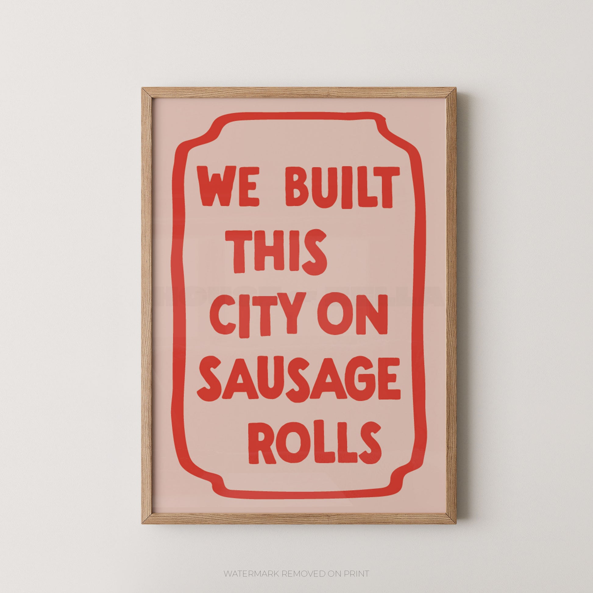 A framed typographic wall art print with a light wood frame, pale pink background, and bold red text reading “WE BUILT THIS CITY ON SAUSAGE ROLLS,” shown hanging against a light neutral wall.