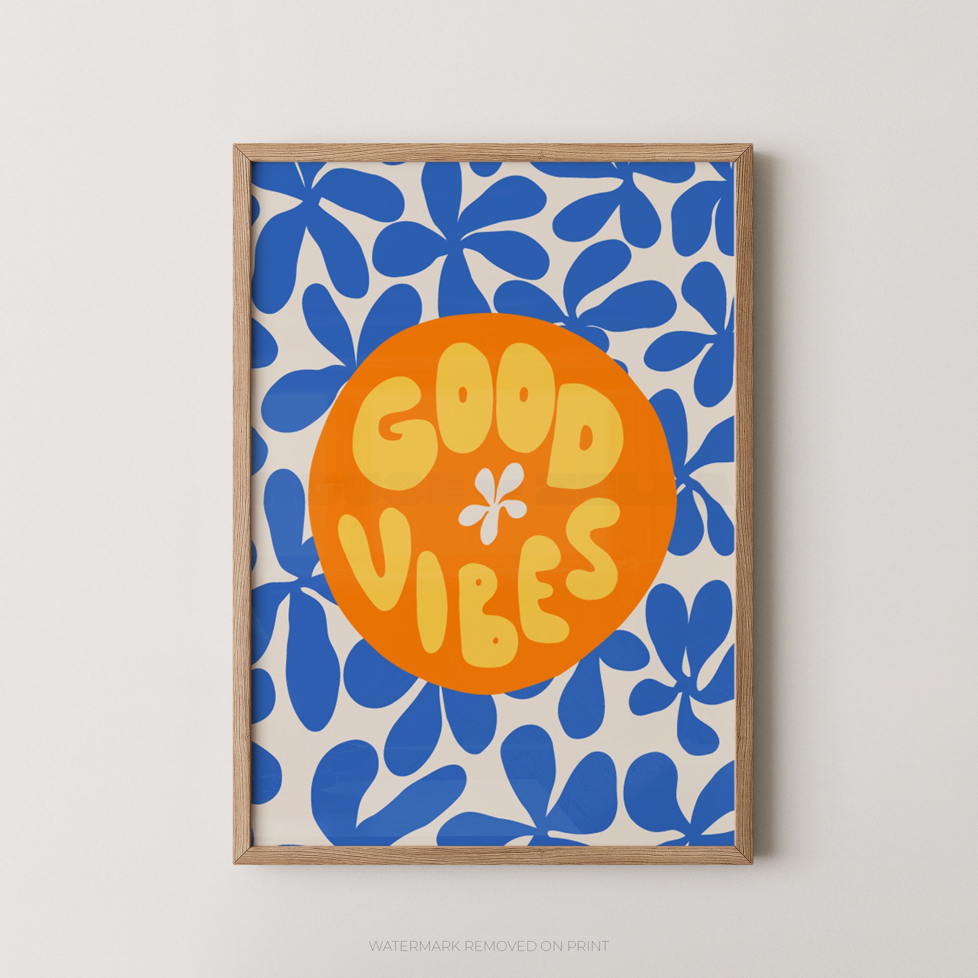 A framed art print featuring a retro pattern of blue flowers on a cream background with a central orange circle containing the text 'GOOD VIBES' in yellow. The print is housed in a light natural wood frame and is displayed against a neutral light gray wall.