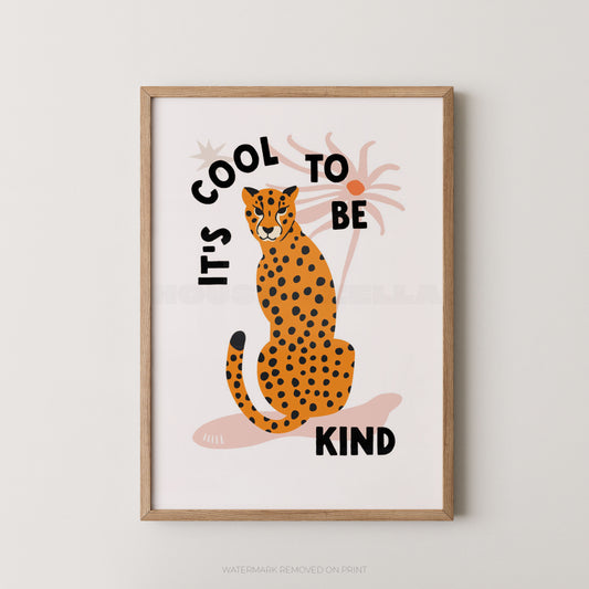 Framed wall art featuring an illustrated orange-and-black spotted cat sitting in front of palm trees, with the text 'IT'S COOL TO BE KIND' in black lettering on a white background.