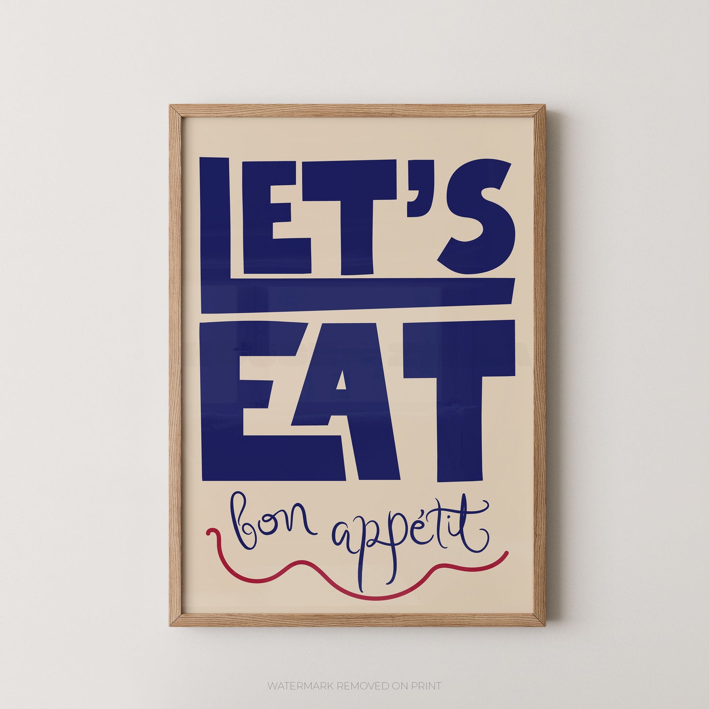Framed wall art print with the phrase “LET’S EAT bon appetit” in bold navy lettering on a warm cream background, with a light natural wood frame, shown against a light gray wall.