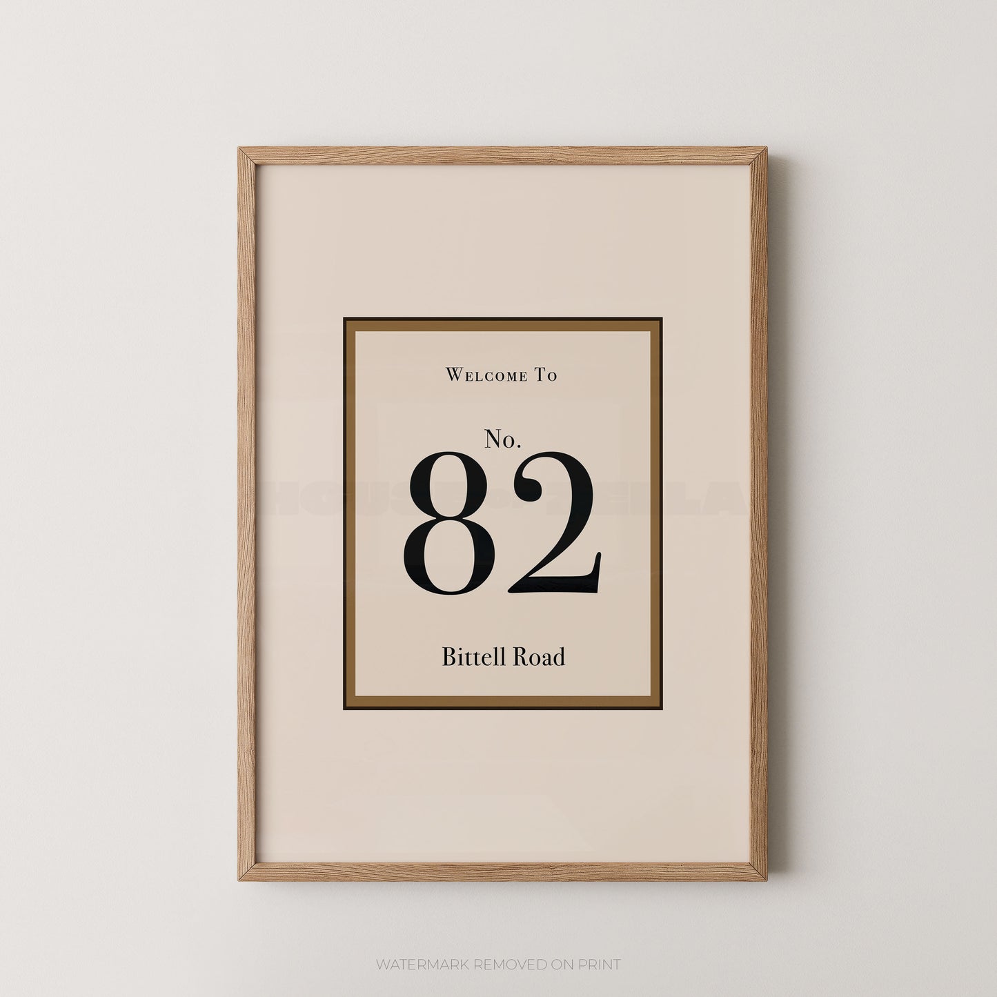 Framed wall art print with a minimalist address design reading 'Welcome To No. 82 Bittell Road' in black serif text on a warm cream background, enclosed in a light natural wood frame against a light gray wall.