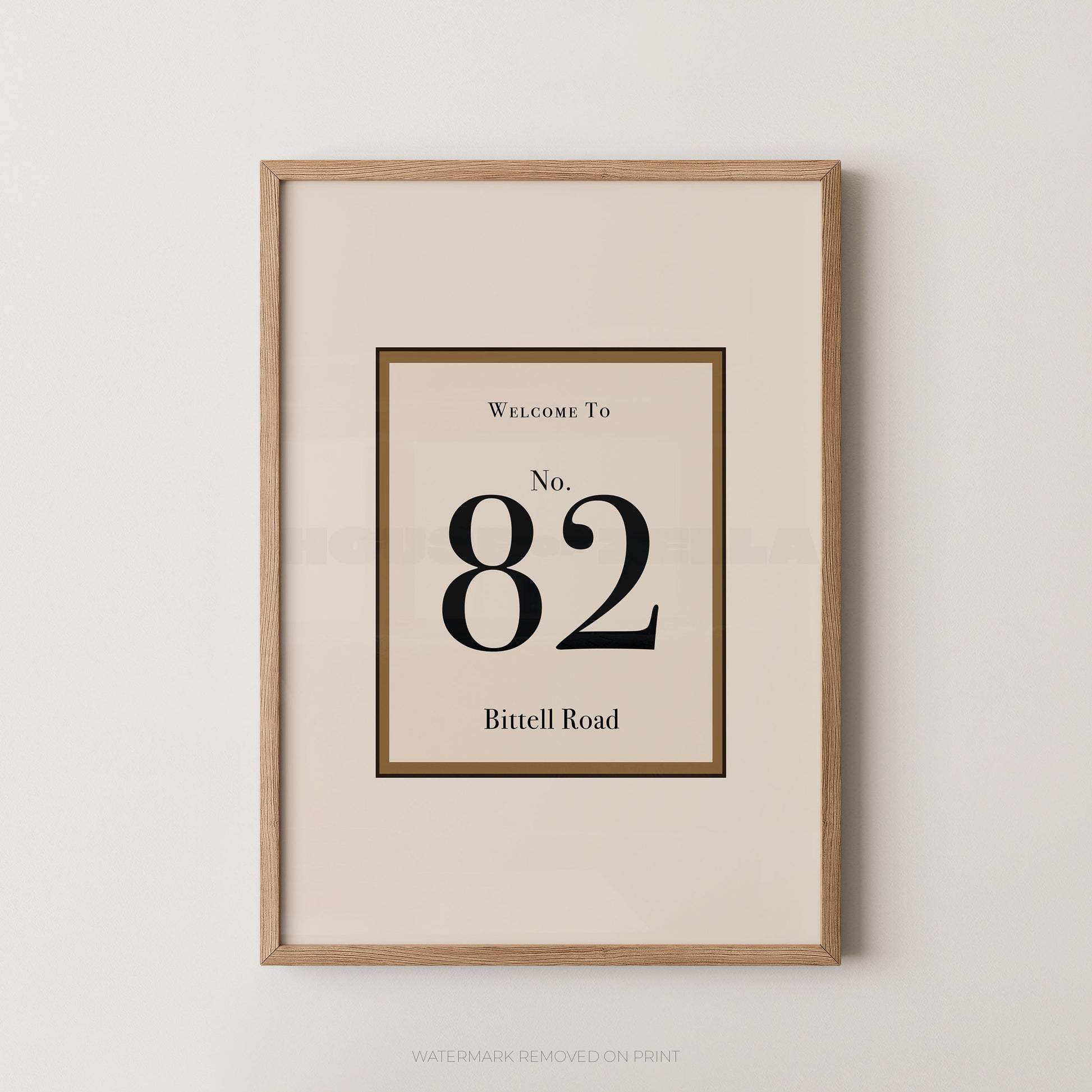 Framed wall art print with a minimalist address design reading 'Welcome To No. 82 Bittell Road' in black serif text on a warm cream background, enclosed in a light natural wood frame against a light gray wall.