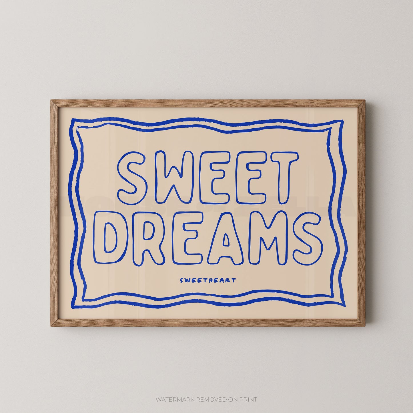 A framed typographic wall art print with the words “SWEET DREAMS” in blue outlined lettering on a warm cream background, enclosed in a light wood frame and shown against a light gray wall.
