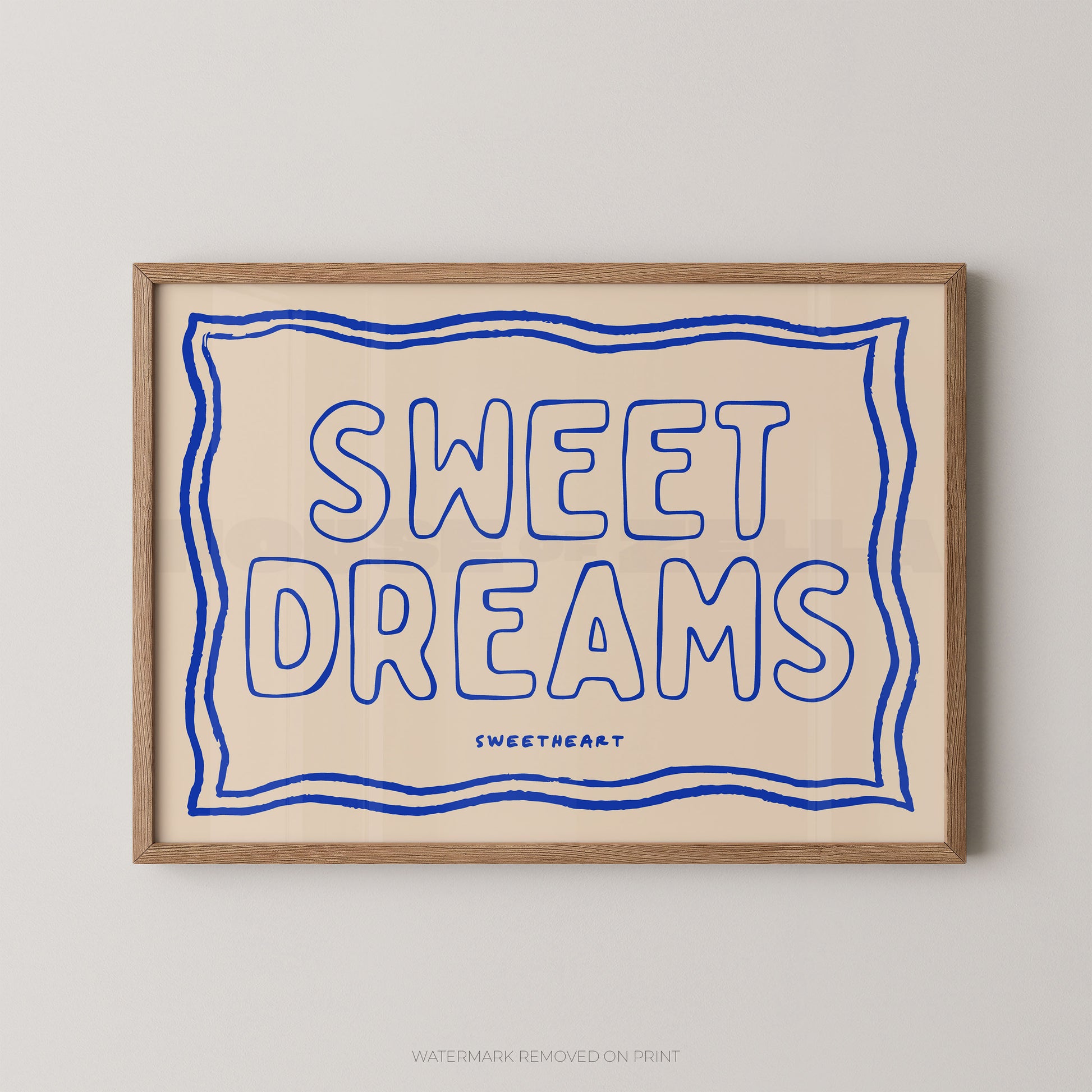 A framed typographic wall art print with the words “SWEET DREAMS” in blue outlined lettering on a warm cream background, enclosed in a light wood frame and shown against a light gray wall.