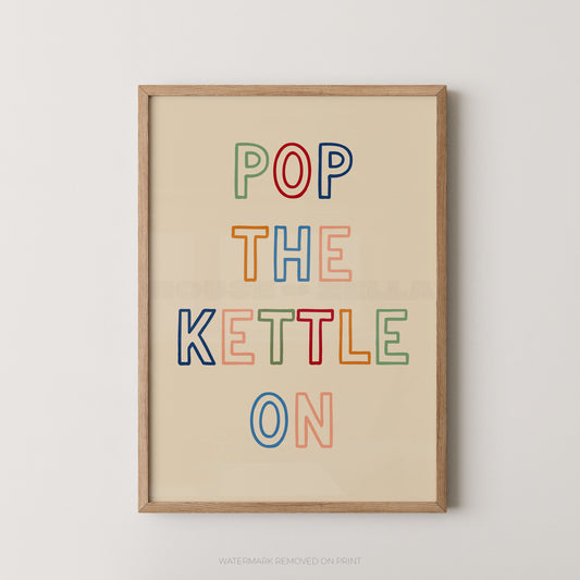 Framed typographic wall art with the phrase “POP THE KETTLE ON” in multicolored outlined letters on a warm cream background, shown hanging against a light neutral wall.