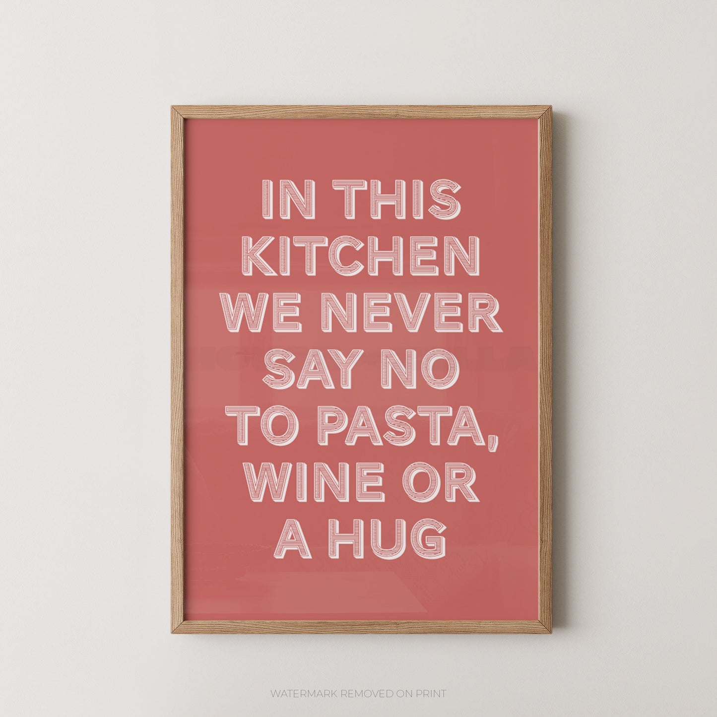 A framed typographic wall art print with a coral pink background and white raised text reading, 'IN THIS KITCHEN WE NEVER SAY NO TO PASTA, WINE OR A HUG,' shown against a light neutral wall.