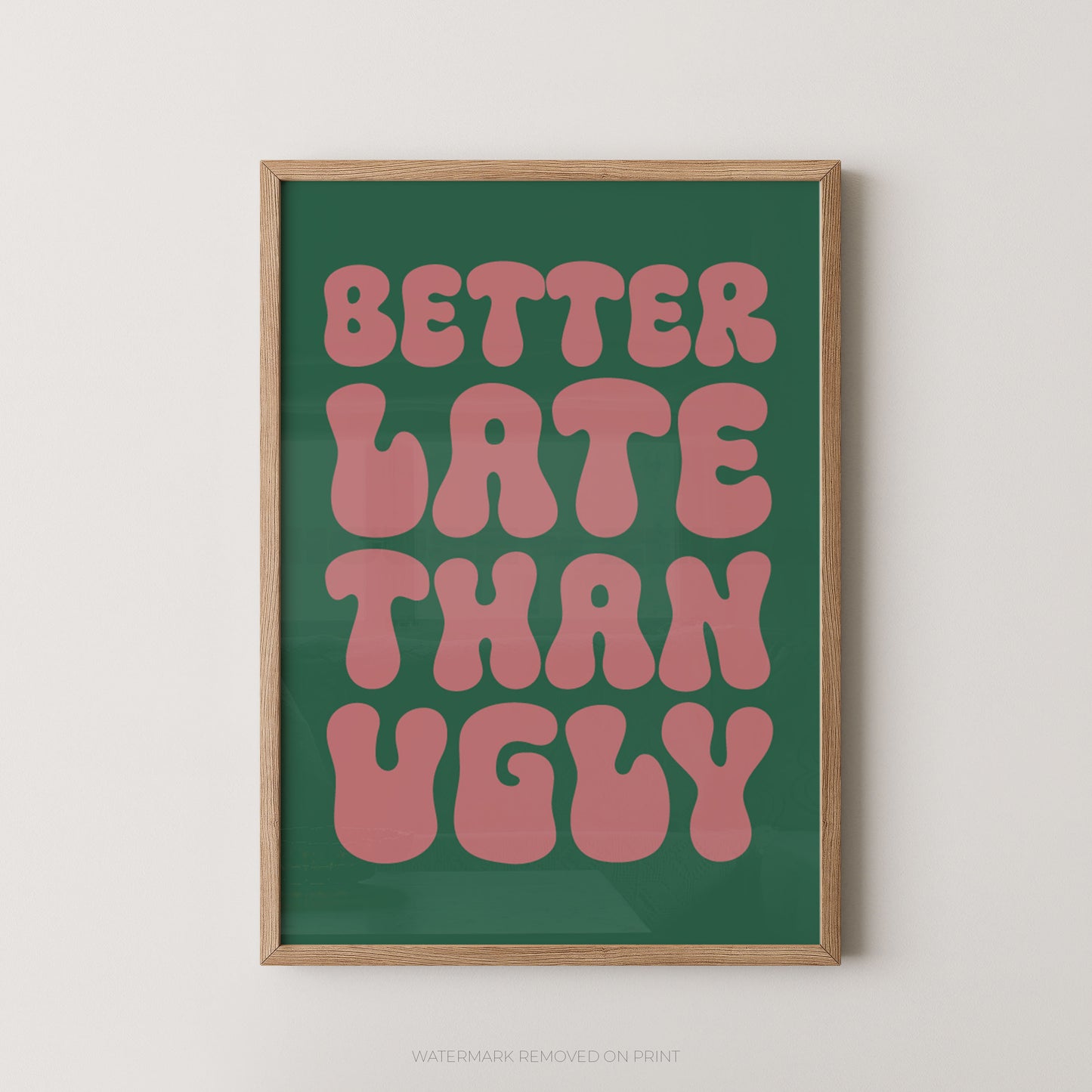 A framed typographic wall art print with the phrase “BETTER LATE THAN UGLY” in pink retro letters on a dark green background, shown in a light wood frame against a plain light wall.