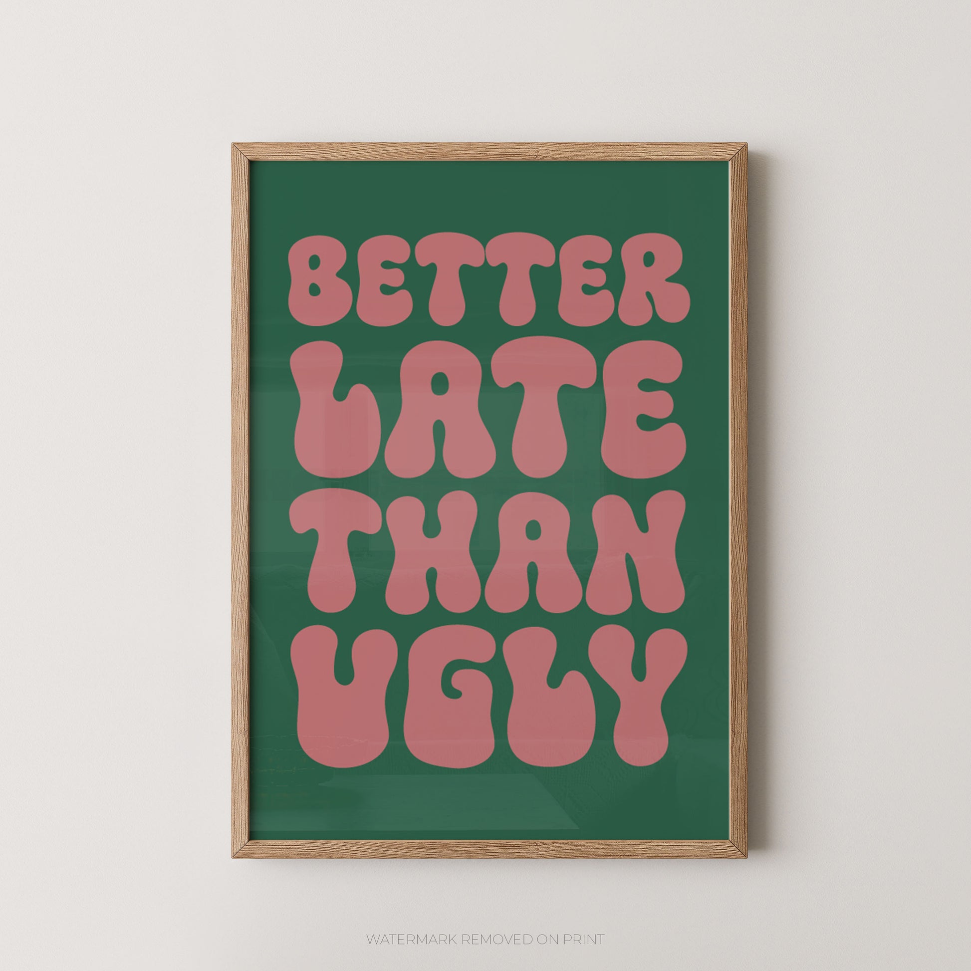 A framed typographic wall art print with the phrase “BETTER LATE THAN UGLY” in pink retro letters on a dark green background, shown in a light wood frame against a plain light wall.