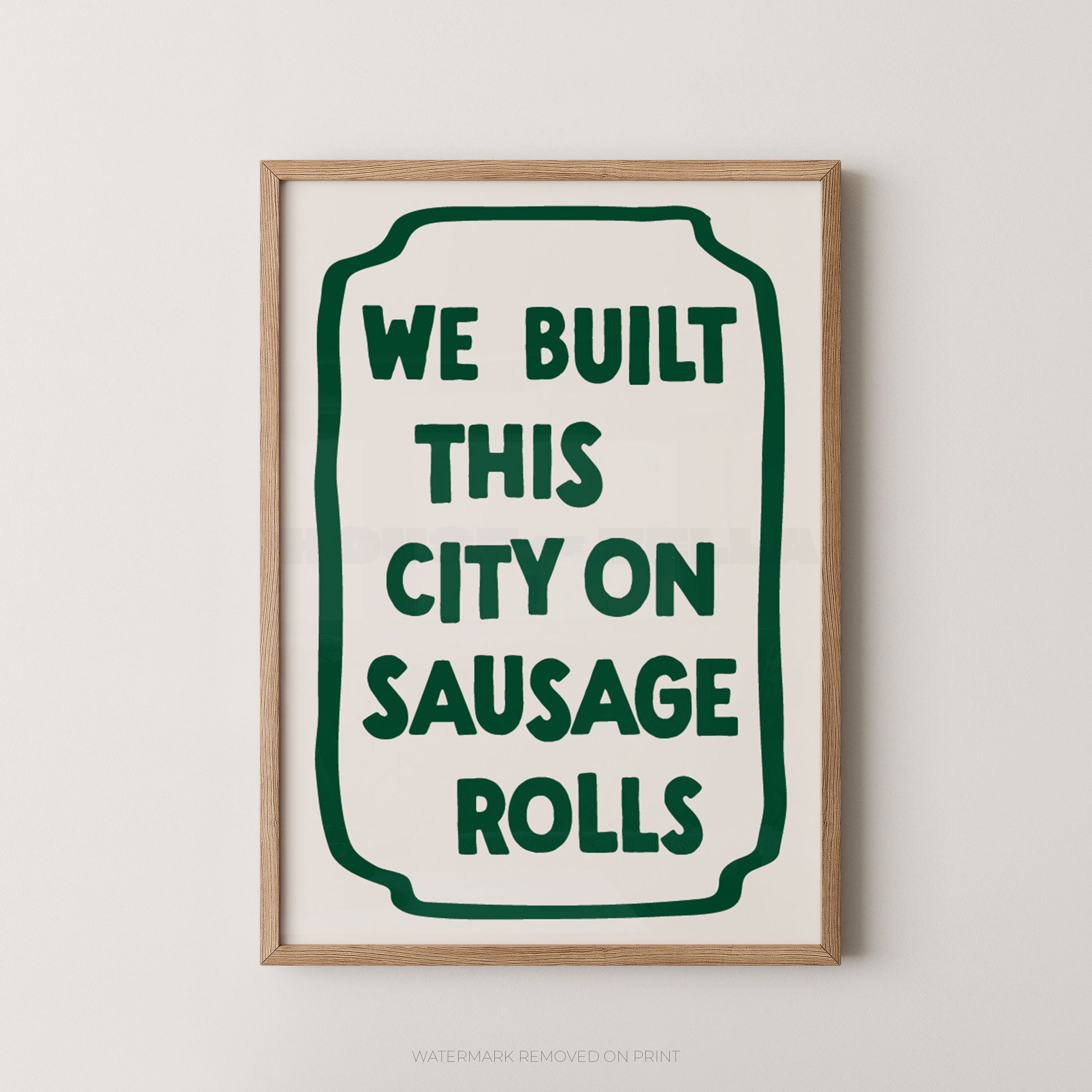A framed typographic wall art print with the phrase “WE BUILT THIS CITY ON SAUSAGE ROLLS” in dark green text inside a rounded green border on an off-white background, shown in a light wood frame against a light gray wall.