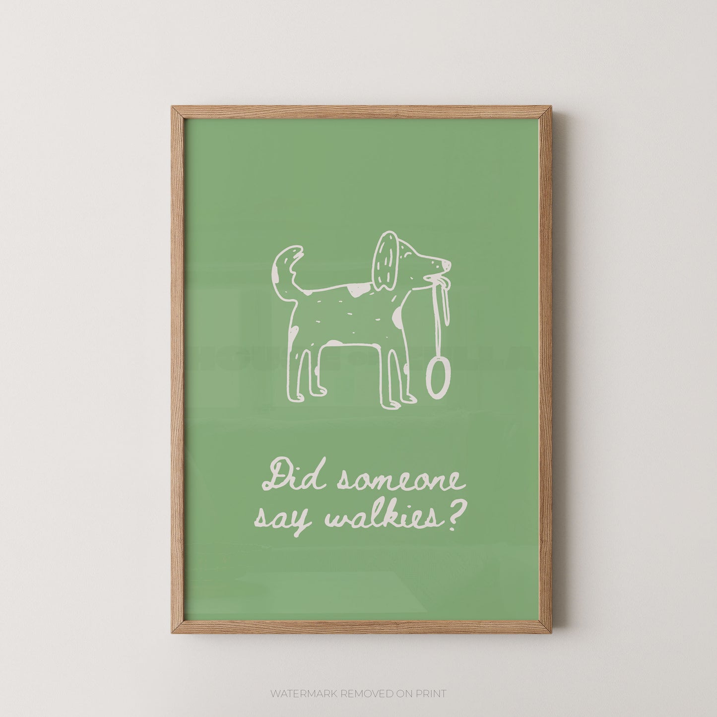 A framed art print featuring a white hand-drawn dog holding a leash on a sage green background, with the text 'Did someone say walkies?' below. The print is housed in a light natural wood frame and is displayed against a neutral light gray wall.