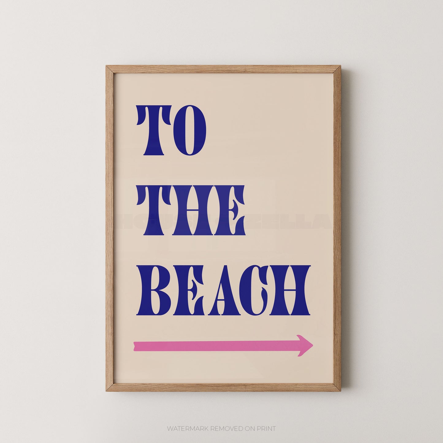 A framed typographic wall art print with the words “TO THE BEACH” in large blue letters on a warm cream background, plus a pink arrow pointing right. The print is shown in a light wood frame against a light neutral wall.