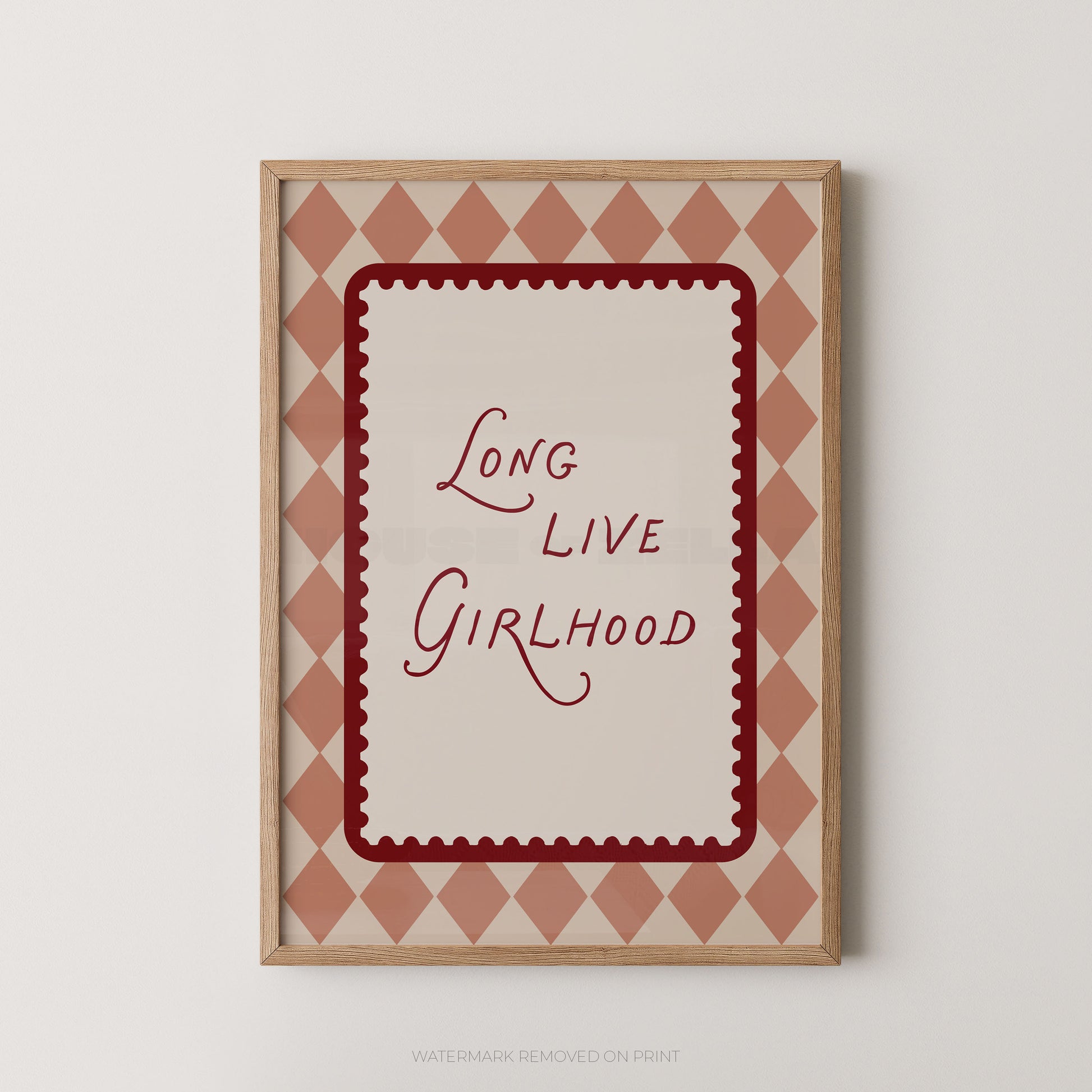 Framed wall art print with the phrase “Long Live Girlhood” in red script, centered on a cream background with a scalloped red border and a dusty pink diamond pattern around the edge. The print is shown in a light wood frame against a plain light gray wall.