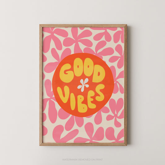 A framed retro-style art print featuring the words 'GOOD VIBES' in yellow inside a large orange circle, set against a pink and cream floral pattern. The print is housed in a light natural wood frame and is displayed on a neutral light gray wall.
