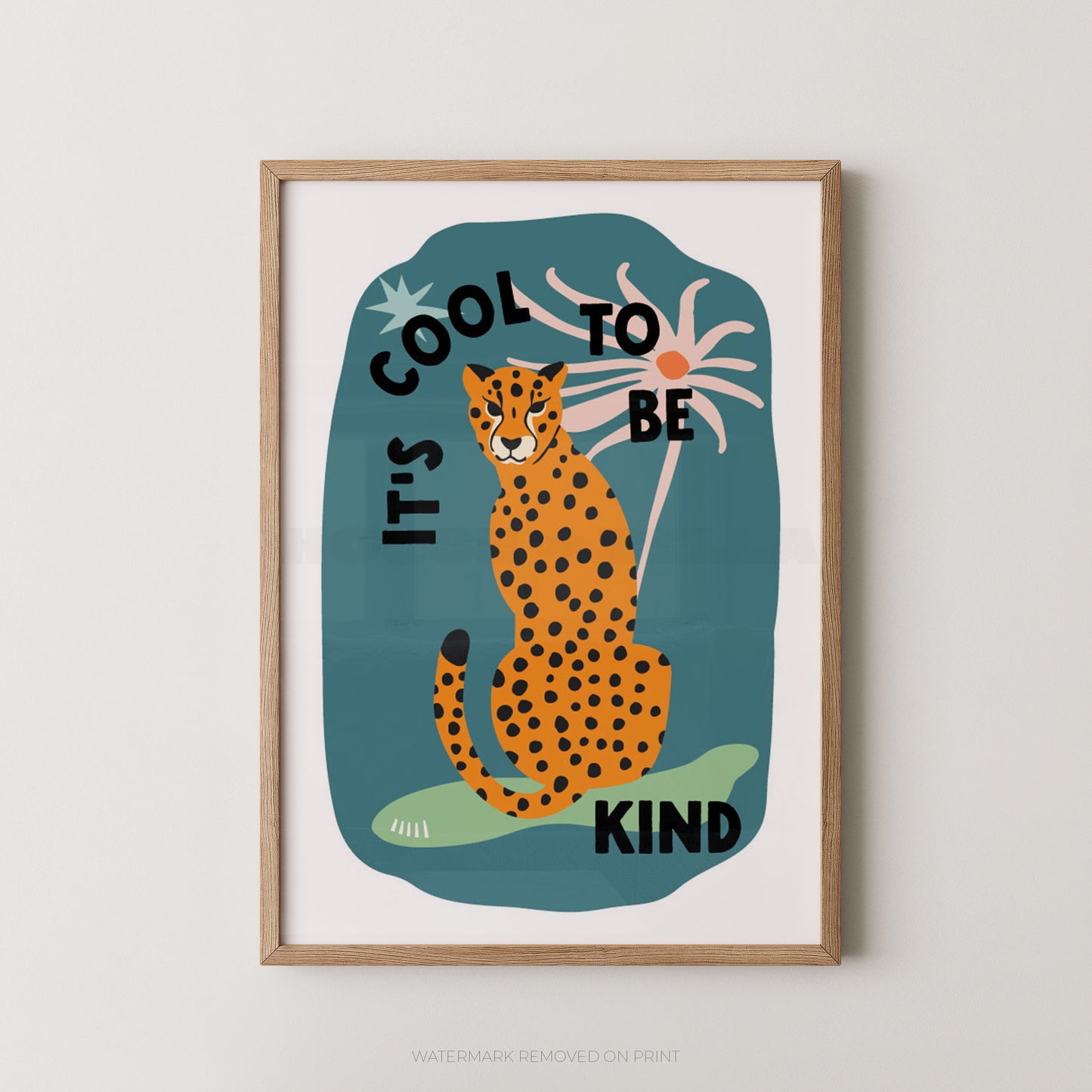 A framed art print featuring a stylized orange-and-black spotted cat sitting on a teal shape with the words “IT’S COOL TO BE KIND” in black lettering, shown against a light neutral wall.