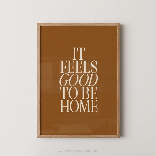 "It Feels Good To Be Home typography wall art print with elegant serif lettering in cream on a warm caramel tan background, displayed straight-on in an oak frame"


