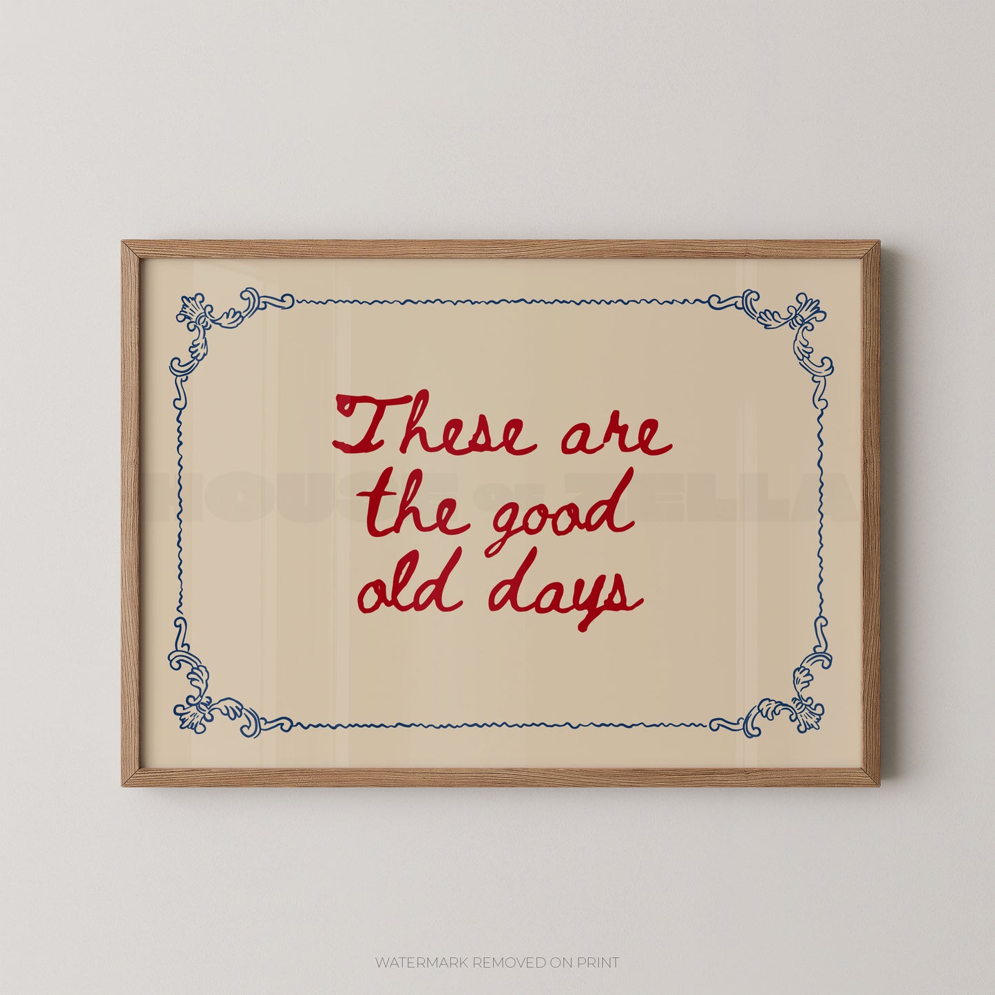 Framed wall art print with the phrase “These are the good old days” in red script, centered on a cream background with a blue decorative border, shown in a light wood frame against a light gray wall.