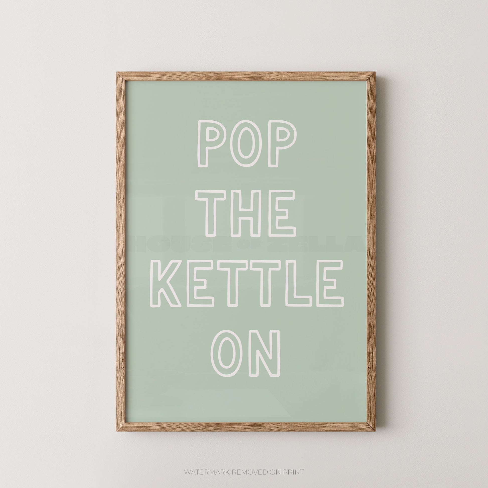 A framed typographic wall art print with the phrase “POP THE KETTLE ON” in white outlined letters on a pale mint green background, shown against a light neutral wall.