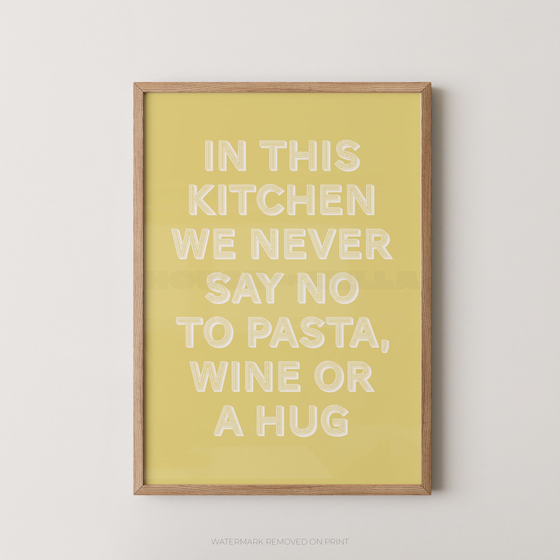 A framed typographic wall art print with a pale yellow background and raised white text reading, 'IN THIS KITCHEN WE NEVER SAY NO TO PASTA, WINE OR A HUG,' shown against a light neutral wall.