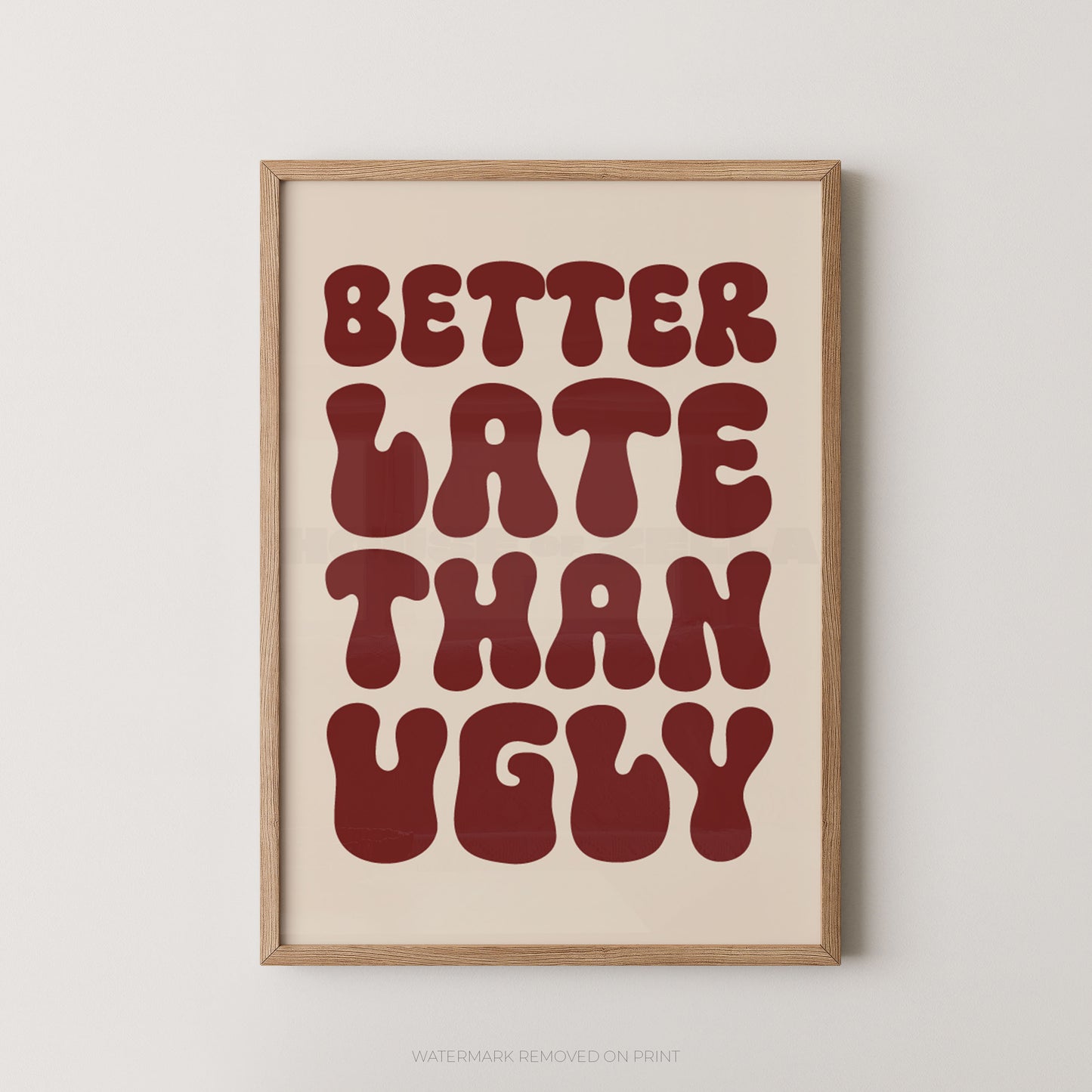 Framed typographic wall art with the phrase “BETTER LATE THAN UGLY” in large dark red retro letters on a warm cream background, shown in a light natural wood frame against a light gray wall.