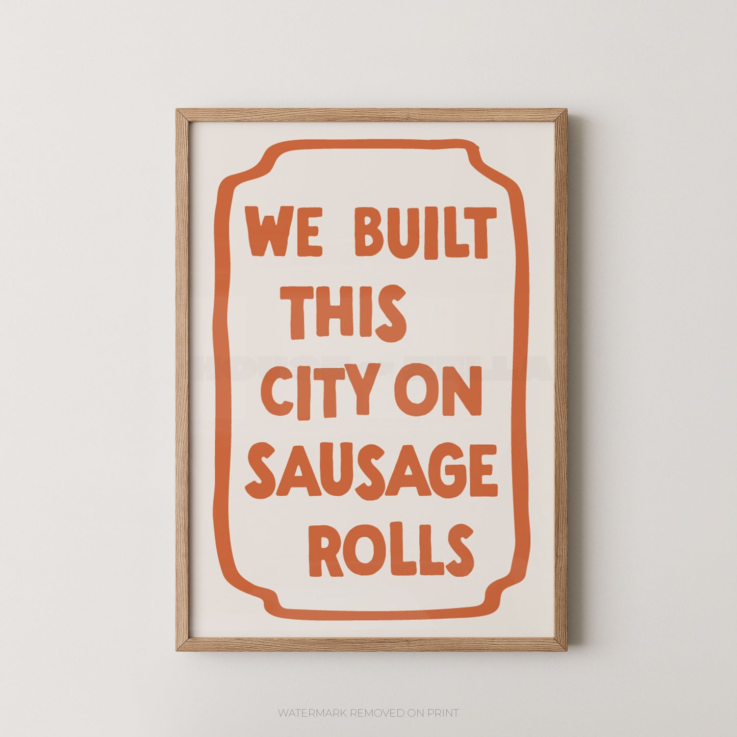 A framed typographic wall art print with the phrase “WE BUILT THIS CITY ON SAUSAGE ROLLS” in orange lettering inside an orange outlined label shape on a white background, shown in a light wood frame against a light gray wall.