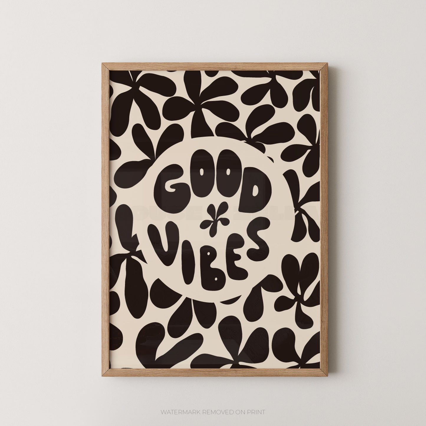 A framed art print featuring a retro pattern of black abstract leaf shapes on a cream background, with a central cream circle containing the text 'GOOD VIBES' in a playful black font. The print is housed in a light natural wood frame and is mounted on a neutral light gray wall.