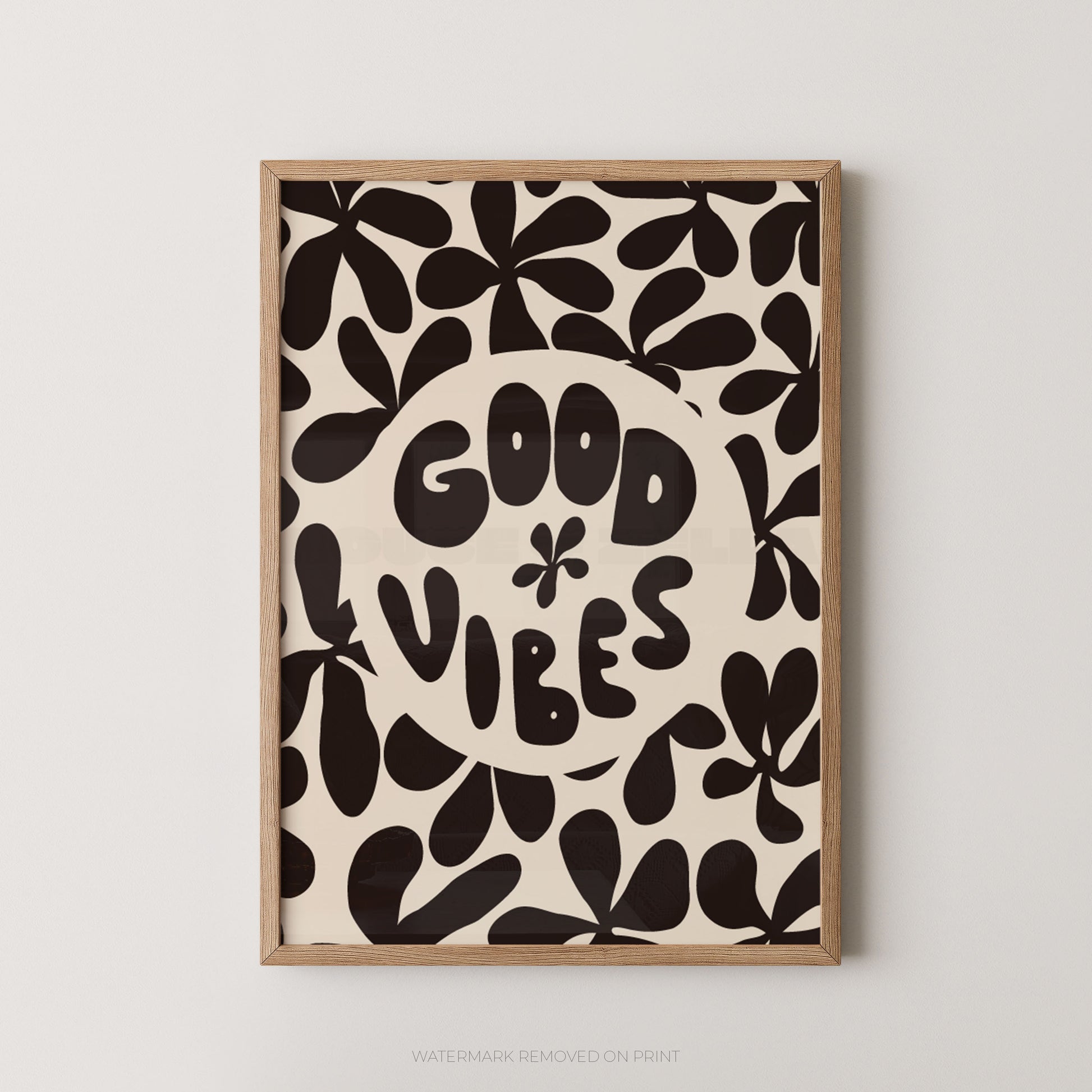 A framed art print featuring a retro pattern of black abstract leaf shapes on a cream background, with a central cream circle containing the text 'GOOD VIBES' in a playful black font. The print is housed in a light natural wood frame and is mounted on a neutral light gray wall.